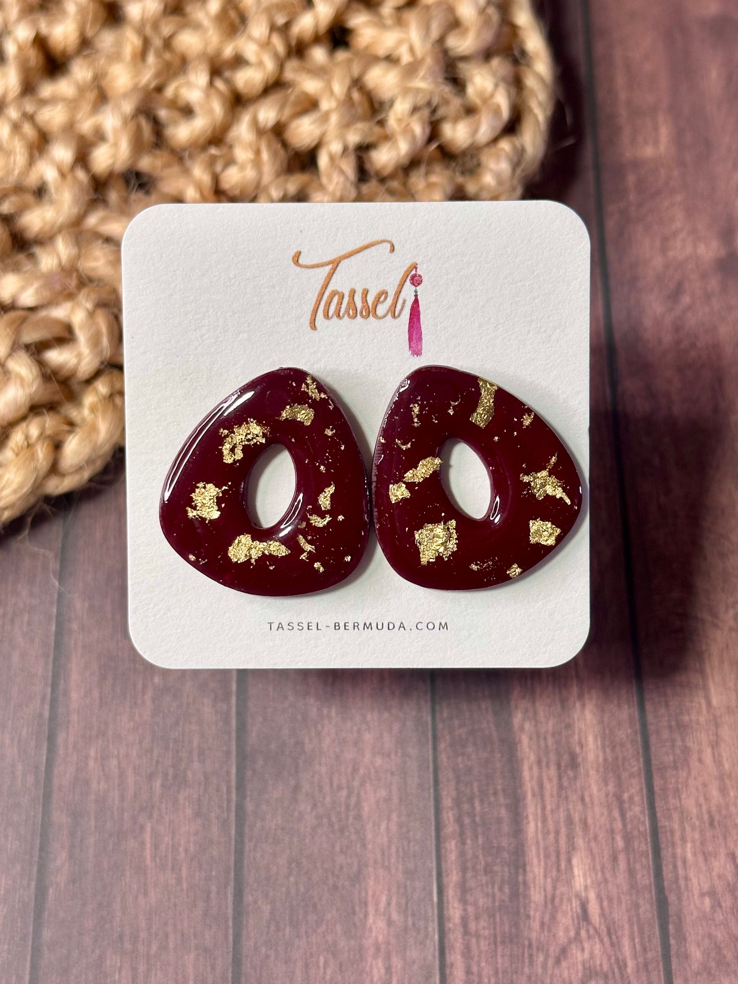 Pair of red stud earrings with gold accents on a Tassel Bermuda branded card, set against a wooden background.