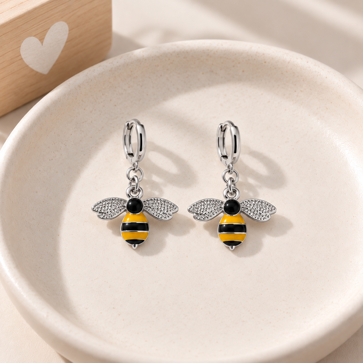 A pair of silver hoop earrings with dangling bee charms, displayed on a light beige ceramic dish. The bee charms feature black and yellow striped bodies and textured silver wings.