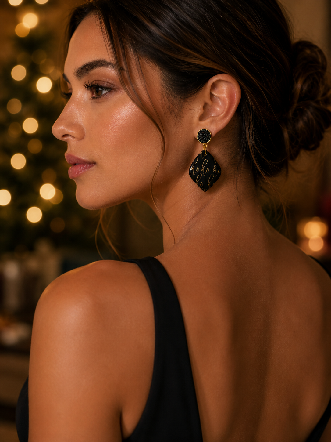 A woman shown from the back and side profile wearing a large black and gold geometric dangle earring by Tassel Bermuda with warm holiday lights in the background.