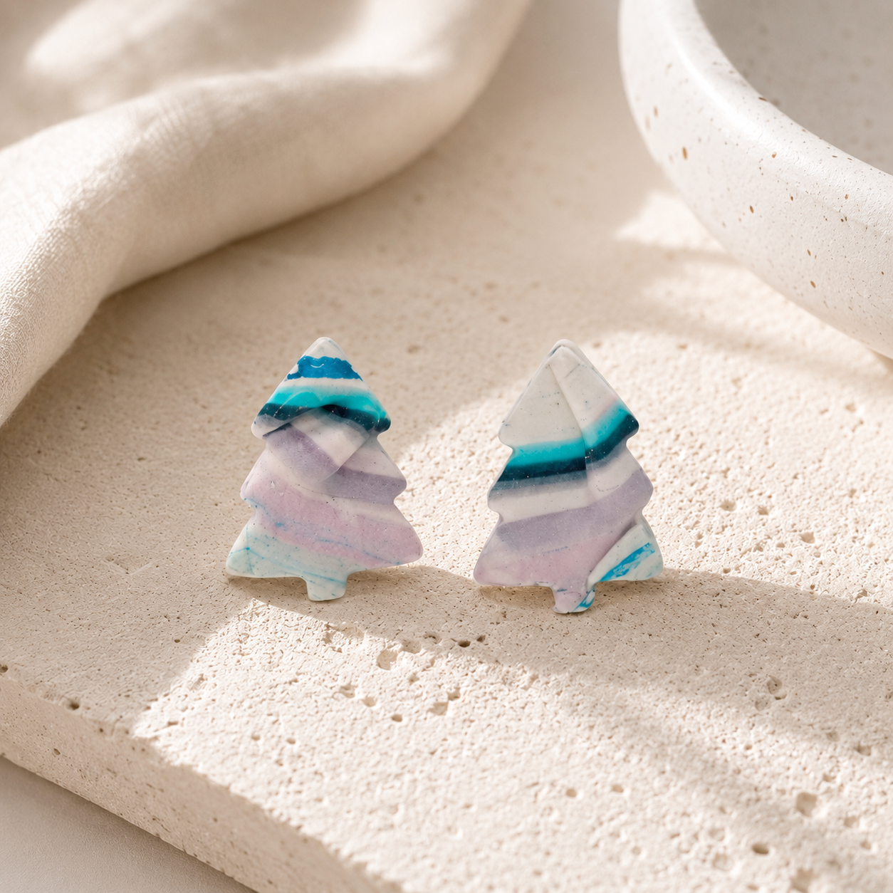 A pair of small tree-shaped stud earrings with marbled pastel layers in white, lavender, light blue, and teal, photographed on a textured beige stone surface with soft natural shadows.