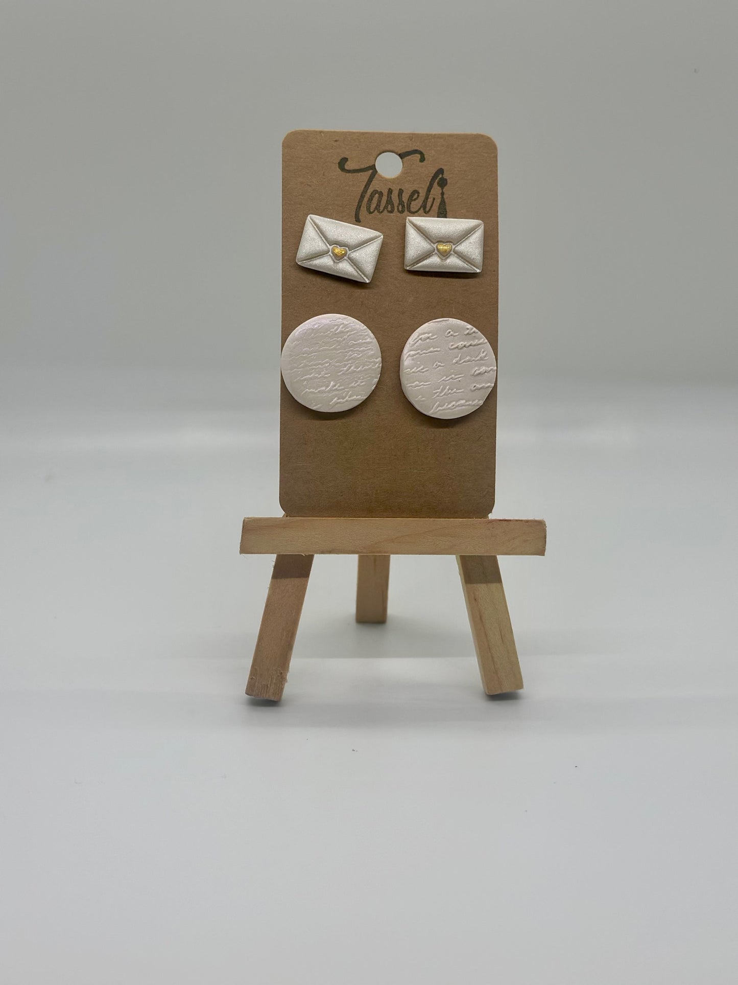 Love Notes polymer clay stud set featuring envelope-shaped earrings and circular handwritten detail studs with shimmering overlay and gold heart accents.
