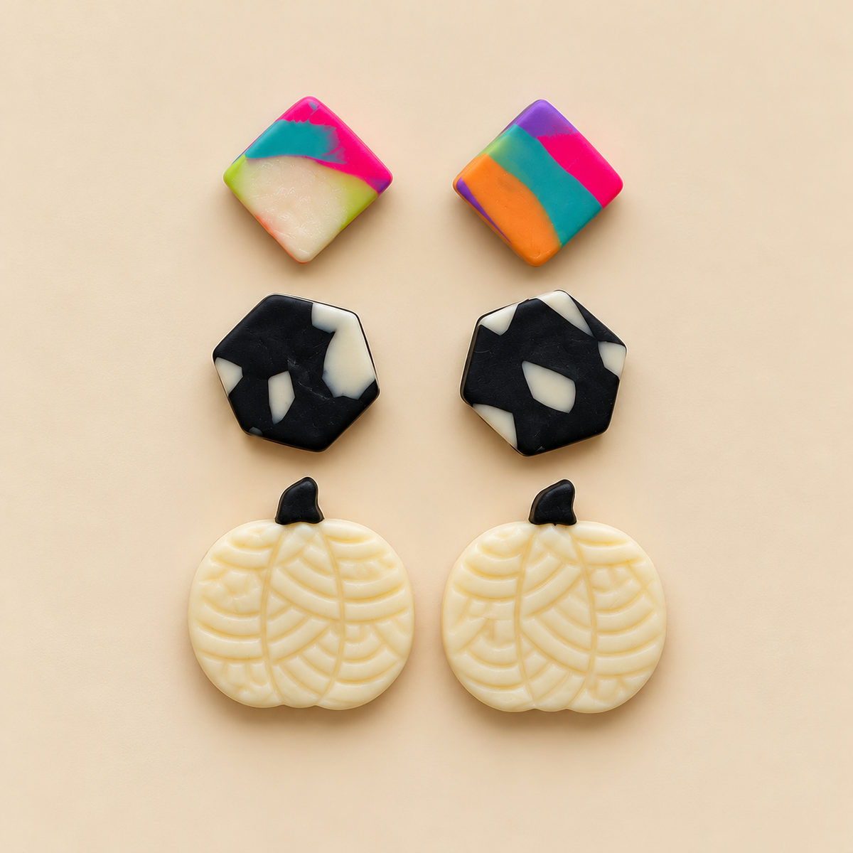 Four decorative jewelry components or charms arranged on a warm beige background: two square pieces with marbled pink, green, yellow, blue, and white patterns, two black-and-white marbled hexagonal pieces, and two cream pumpkin-shaped pieces with black stem tops and leaf-like patterning.