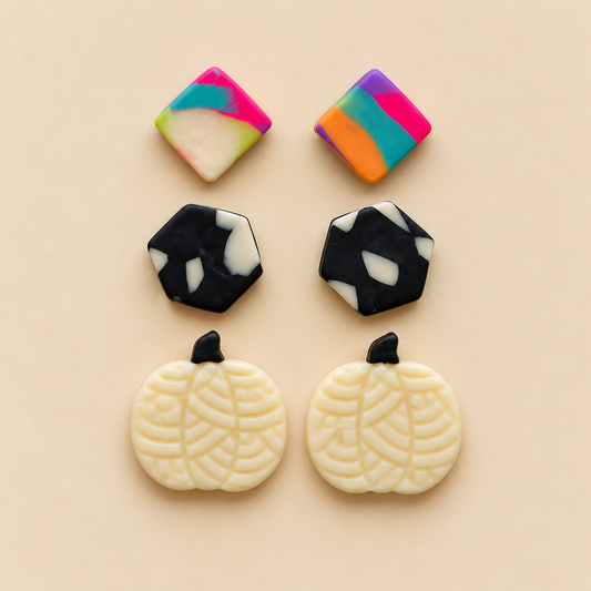Four decorative jewelry components or charms arranged on a warm beige background: two square pieces with marbled pink, green, yellow, blue, and white patterns, two black-and-white marbled hexagonal pieces, and two cream pumpkin-shaped pieces with black stem tops and leaf-like patterning.