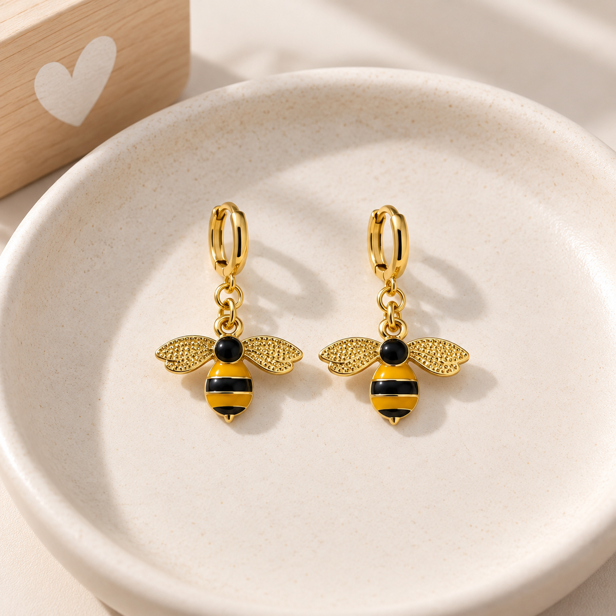 A pair of gold hoop earrings with dangling bee charms, displayed on a round beige ceramic dish.
