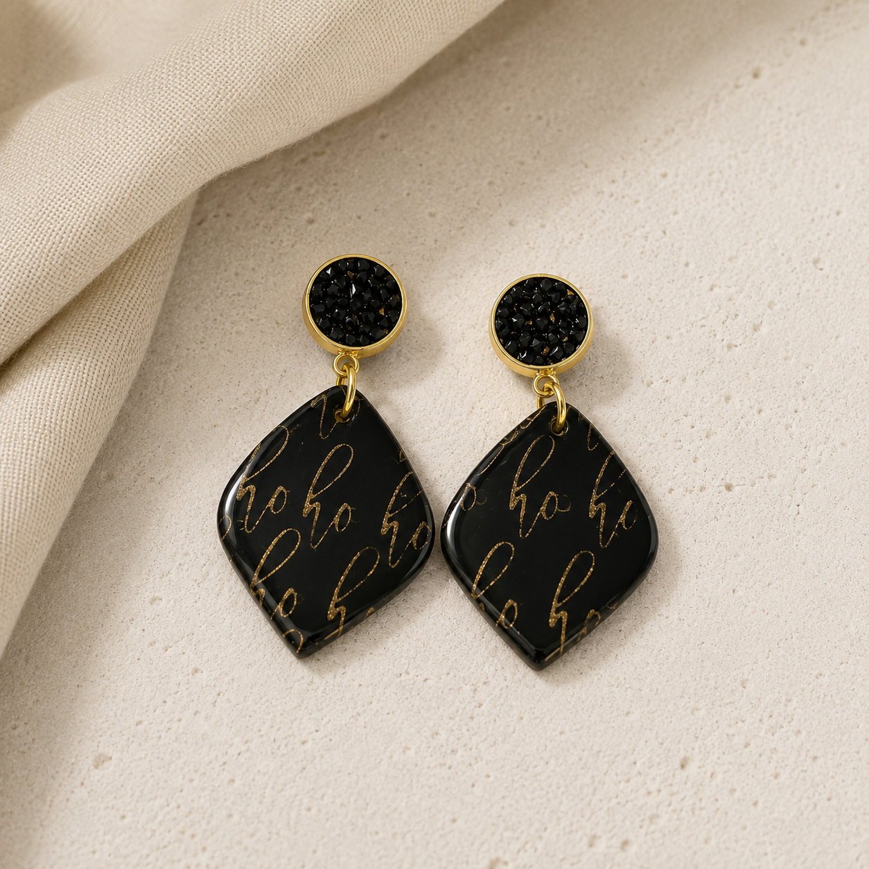 A pair of black shimmery stud earrings with teardrop-shaped drop pendants featuring gold foil 'ho ho' script, displayed on a textured beige surface with a draped cream fabric.