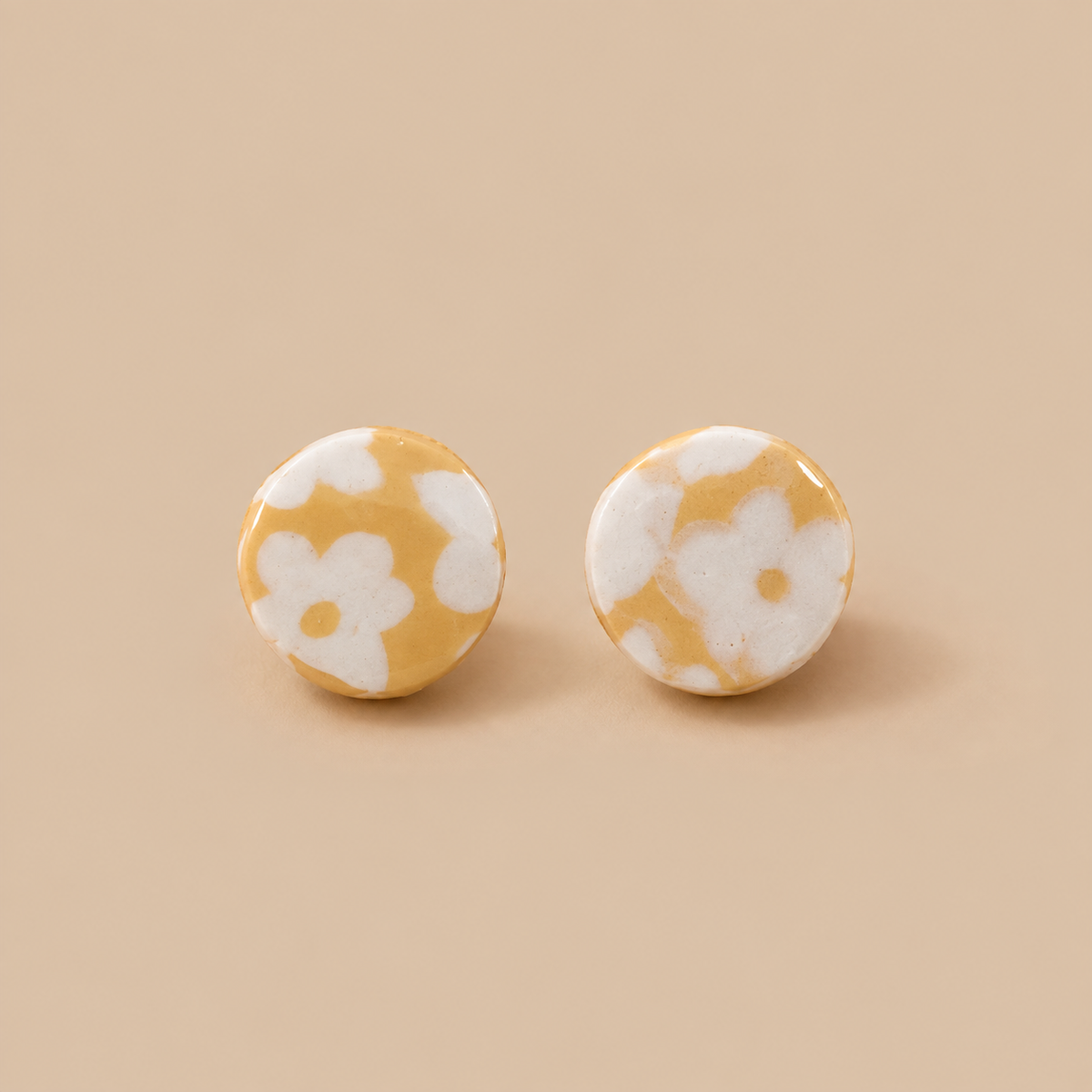 A pair of round stud earrings with a yellow and white floral pattern, displayed on a solid beige background.