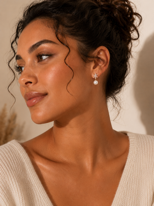 A woman with dark hair in a bun wearing a silver-toned starburst stud earring with a white pearl drop, shown in a warm, close-up portrait against a beige background.