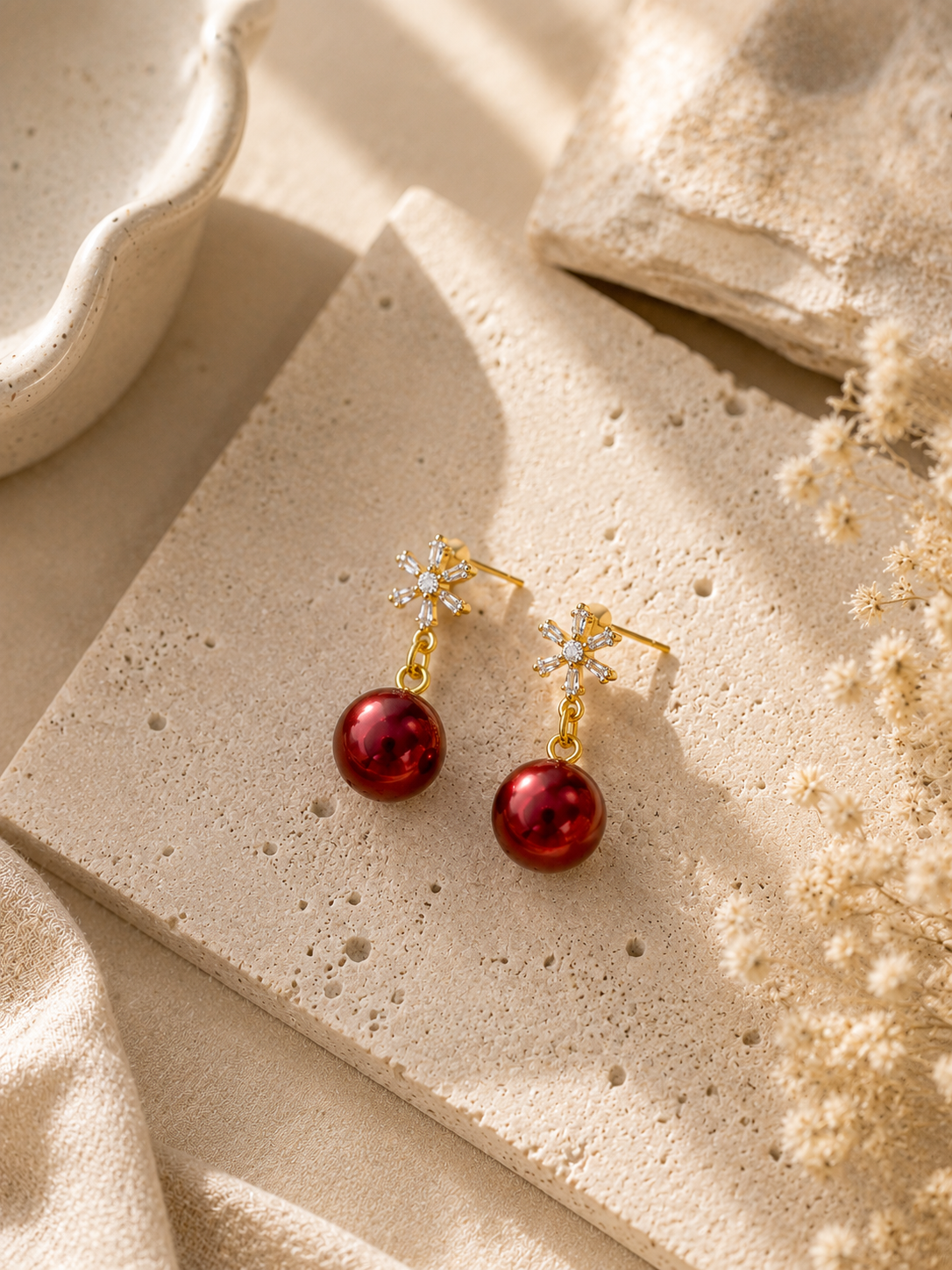 A pair of gold drop earrings with small clear crystal snowflake details and round deep red pearl bead, displayed on a textured beige stone tile with neutral props.