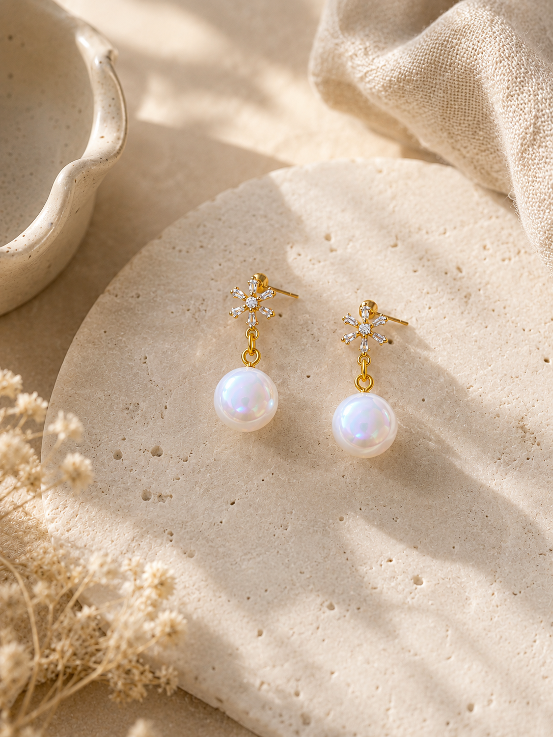 A pair of gold dangle earrings with small clear crystal snowflake  and a single white pearl drop, displayed on a textured beige stone slab with dried flowers and neutral fabric in the background.