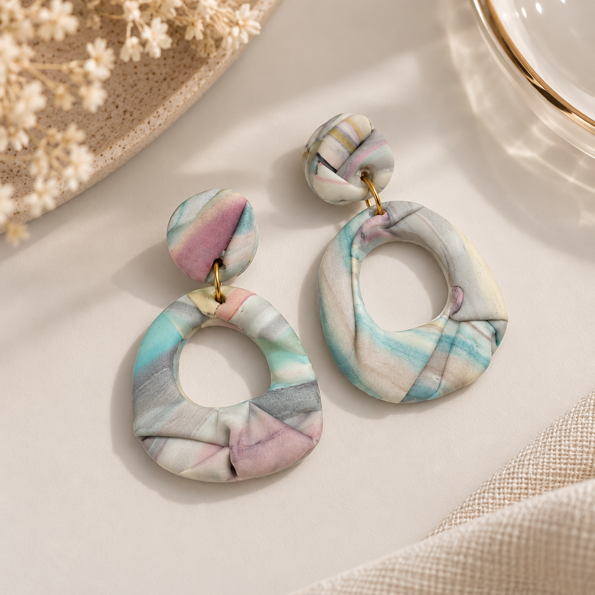 A pair of large, circular dangle earrings made from draped polymer clay with a pastel marbled pattern in pink, blue, green, and gray, hanging from gold-tone rings on a light beige surface.
