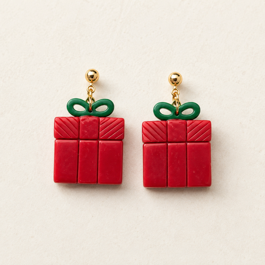A pair of red gift box earrings with green ribbon bows and gold-tone posts, displayed on a light beige textured background.