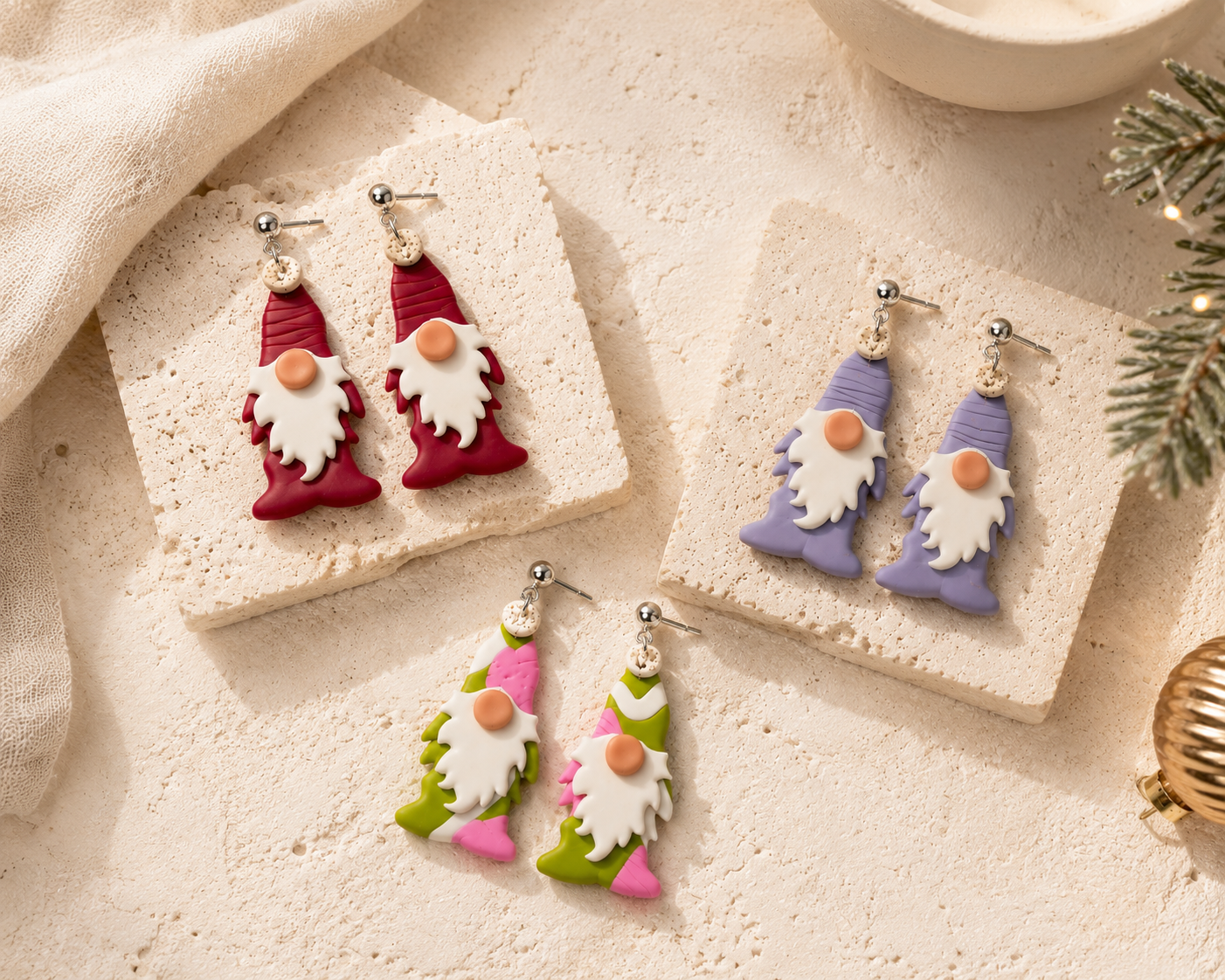 Three pairs of gnome-shaped dangle earrings displayed on textured stone tiles and fabric. The earrings feature fish-shaped bodies in burgundy, lavender, and green, with white beards, peach faces, and silver fishhook ear wires.