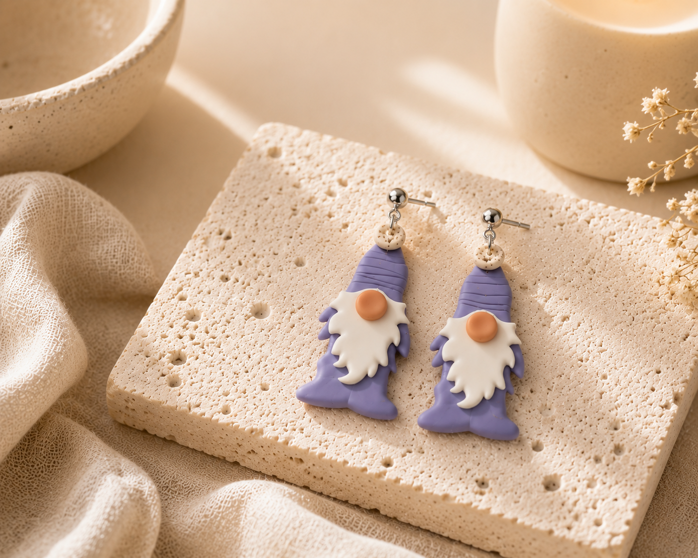 A pair of dangling gnome earrings with lavender-purple hats and boots, white beards, and orange noses, displayed on a textured beige stone slab with neutral fabric and ceramic props.