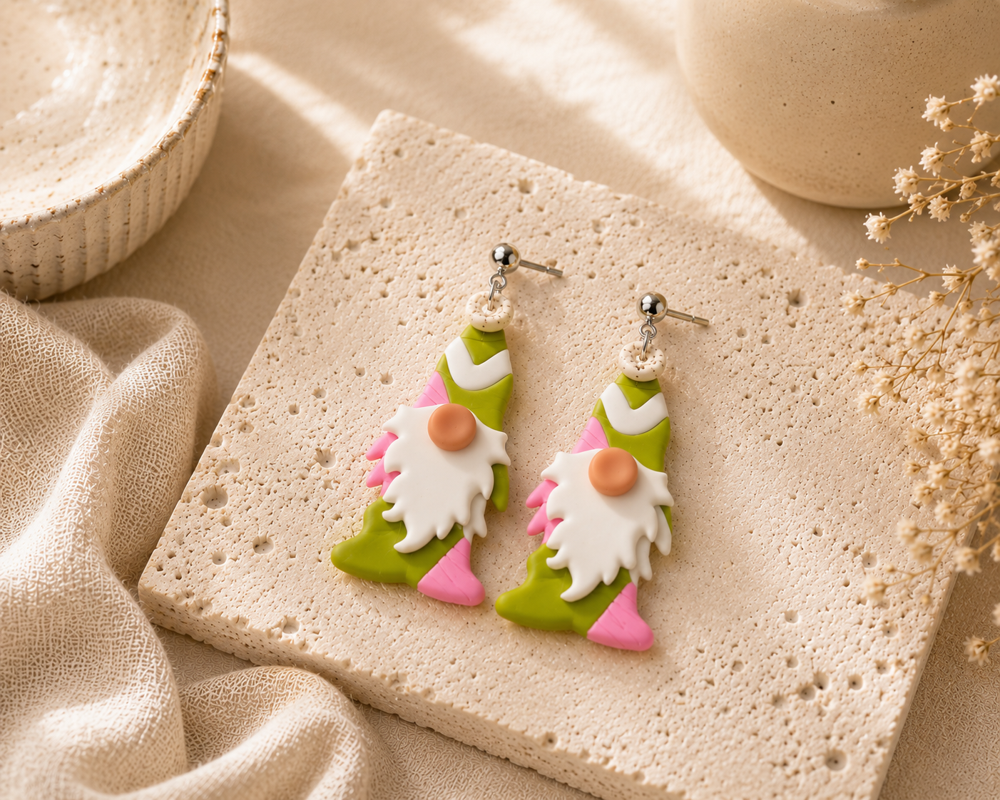 A pair of handmade polymer clay earrings shaped like gnomes, displayed on a textured beige stone slab with neutral fabric and dried floral props.