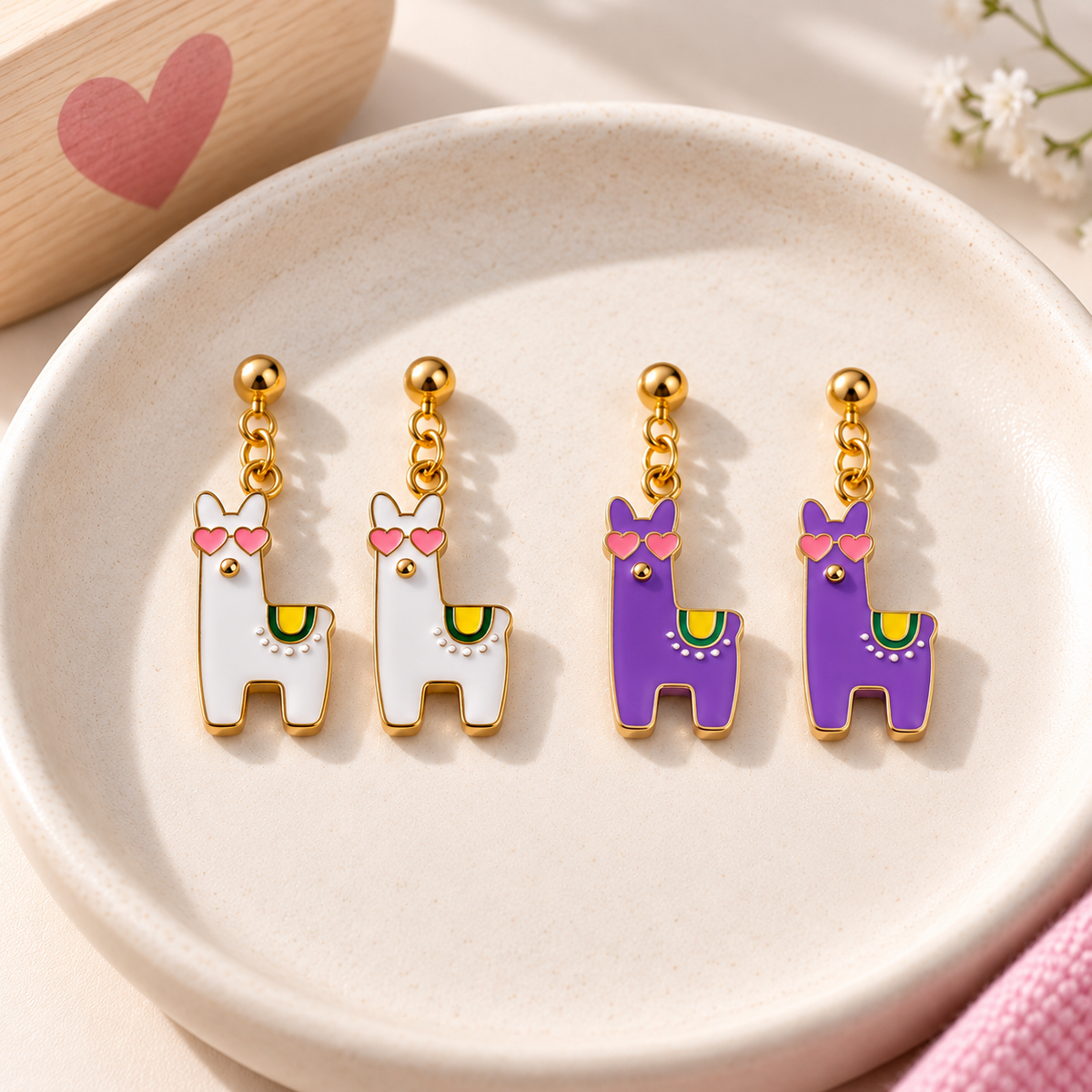 Four llama-shaped dangle earrings displayed on a light beige ceramic dish. Two earrings are white with pink heart sunglasses and yellow-green accents, and two are purple with pink bow ties and yellow-green accents.