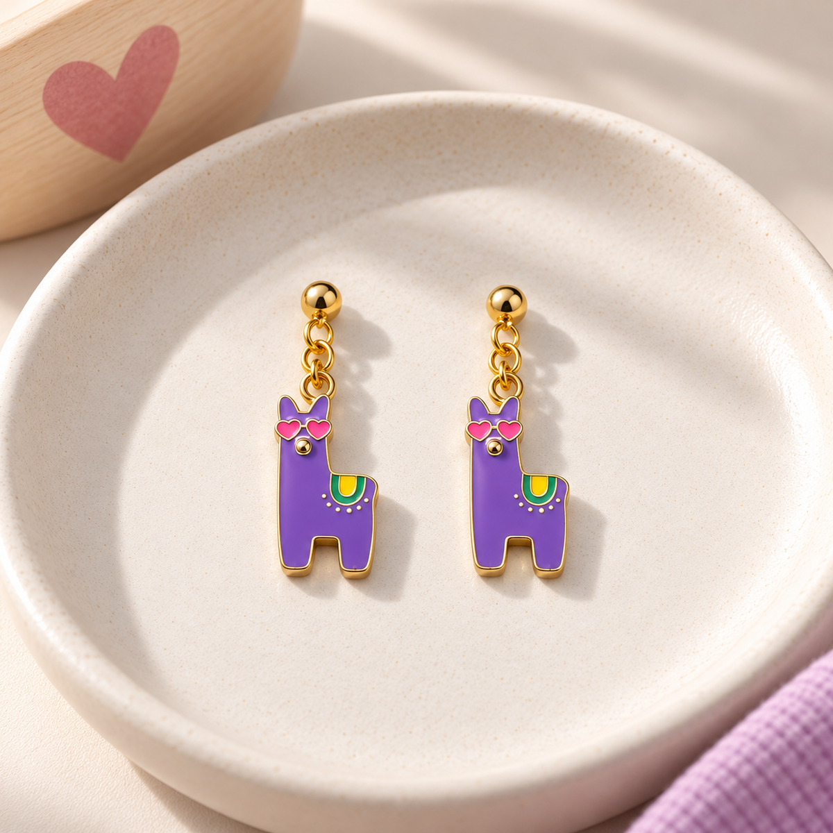 A pair of dangling llama-shaped earrings in purple enamel with gold-tone metal, pink sunglasses, and small multicolor accents, displayed on a round white dish.