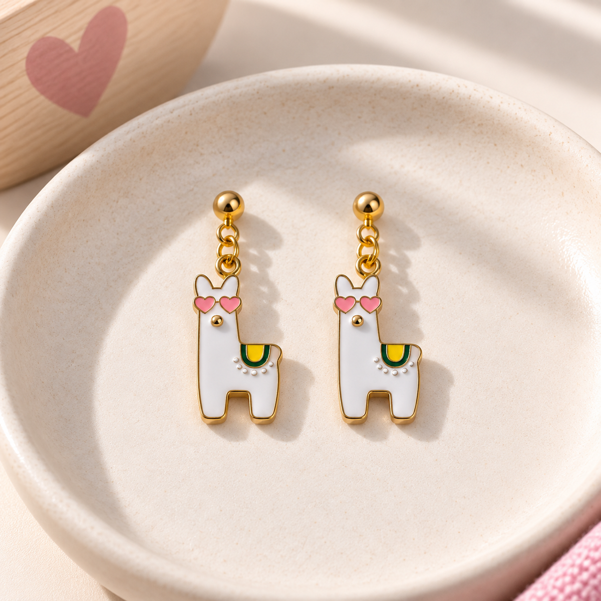 A pair of gold-tone dangle earrings shaped like white llamas with pink heart-shaped sunglasses and a small green accent, displayed on a round beige dish.