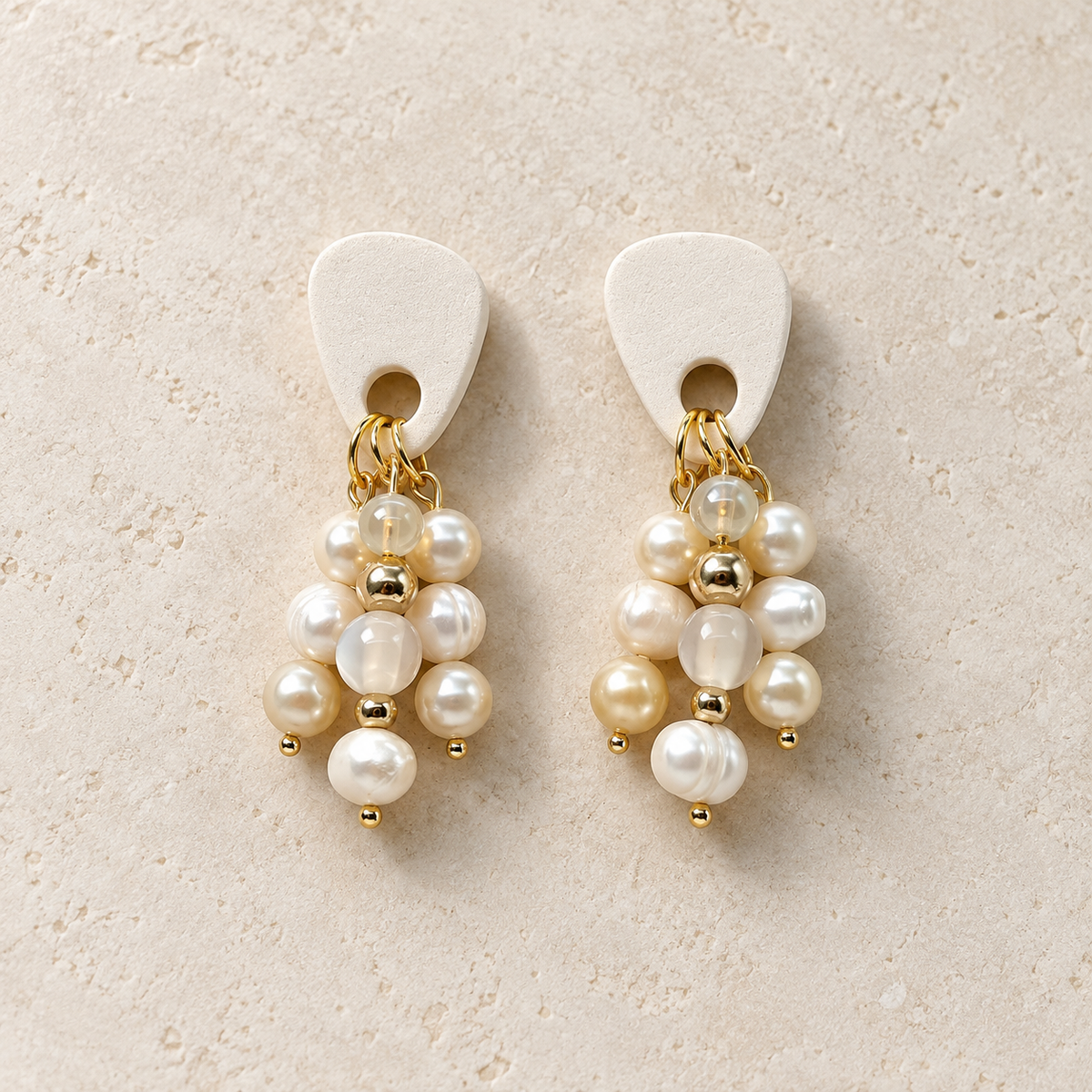 A pair of drop earrings with off-white guitar pick-shaped tops, gold-tone chains, and clusters of white and cream pearls with small gold beads, displayed on a textured beige surface.