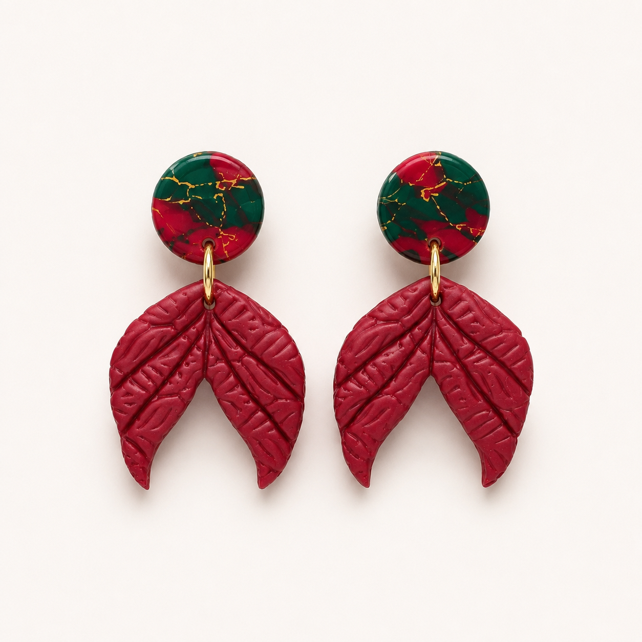A pair of statement dangle earrings featuring textured deep red leaf-shaped drops and circular studs with a red and green marbled pattern accented by gold veins, shown on a clean off-white background.