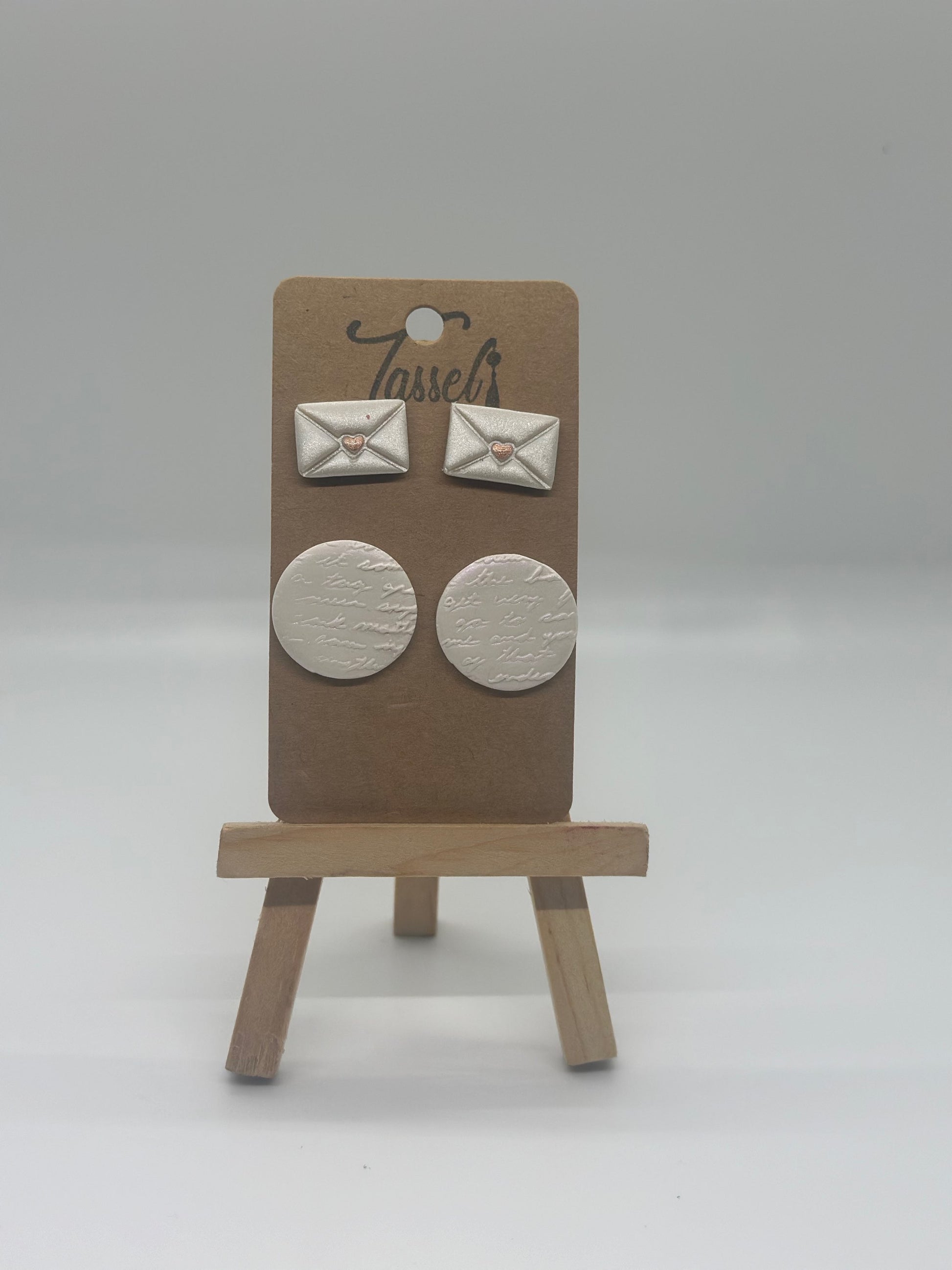 Love Notes polymer clay stud set featuring envelope-shaped earrings and circular handwritten detail studs with shimmering overlay and gold heart accents.