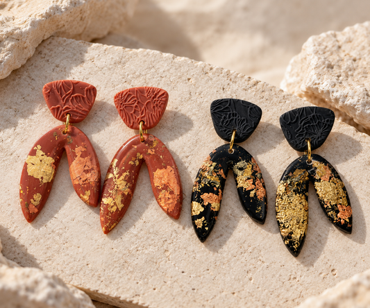 Four dangle earrings displayed on a light beige stone surface outdoors. Two earrings are terracotta red with gold leaf inlay, and two are black with gold leaf inlay.