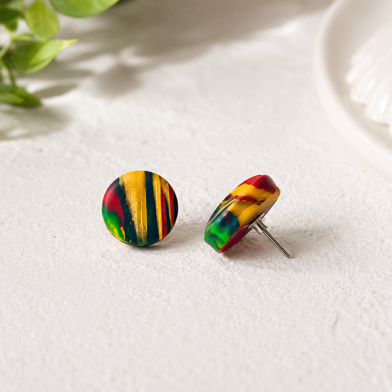A pair of round stud earrings with a colorful, marbled pattern of yellow, red, green, and black, displayed on a textured white surface with soft shadows.