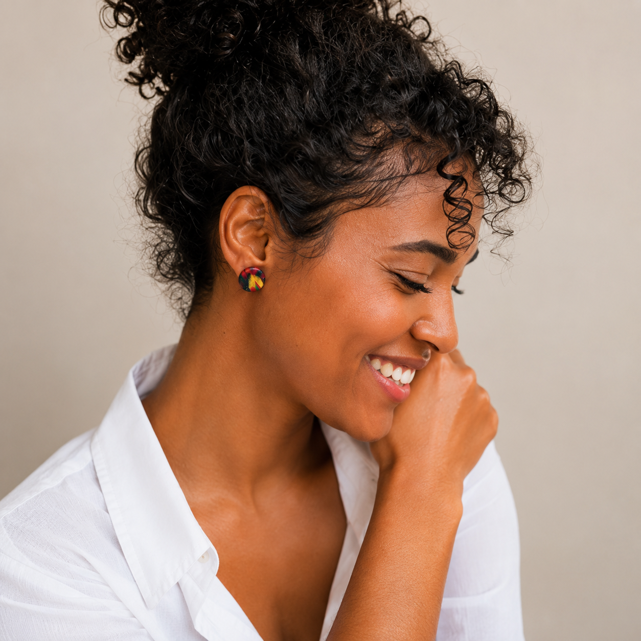 A woman with curly black hair wearing a white collared shirt and small round earrings with a yellow and black pattern, posing against a neutral beige background.