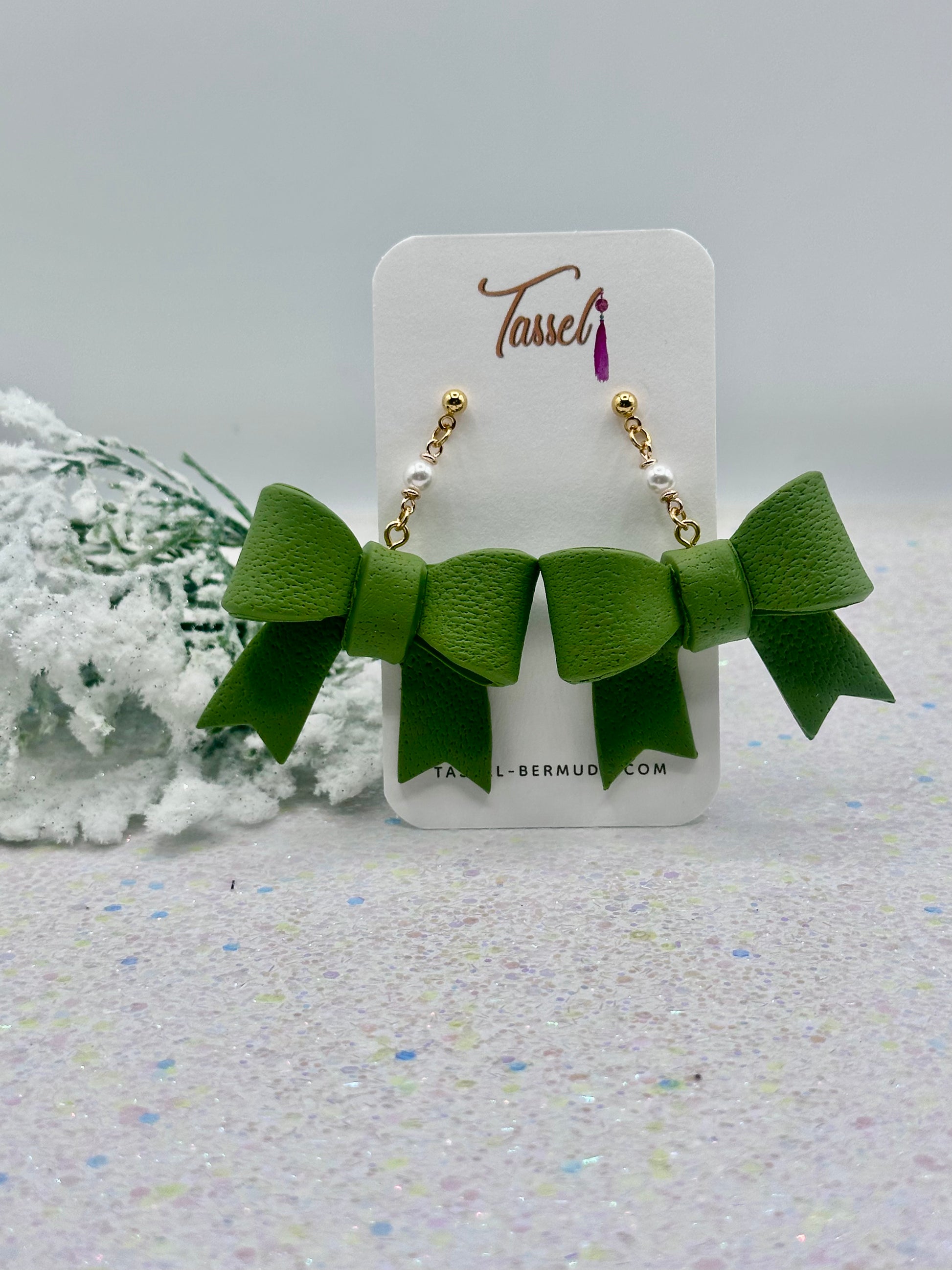 Marie Green dangle earrings with white polymer clay bows and pearl-accented chain displayed on Tassel Bermuda card with snowy holiday backdrop.