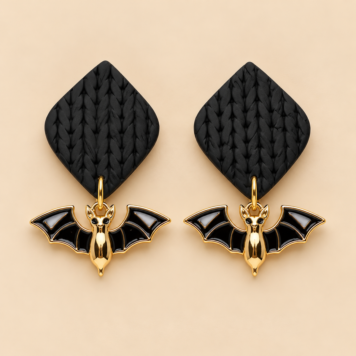 A pair of gothic-style bat earrings with textured black diamond-shaped studs and gold bat charms with black enamel wings, displayed on a warm beige background.