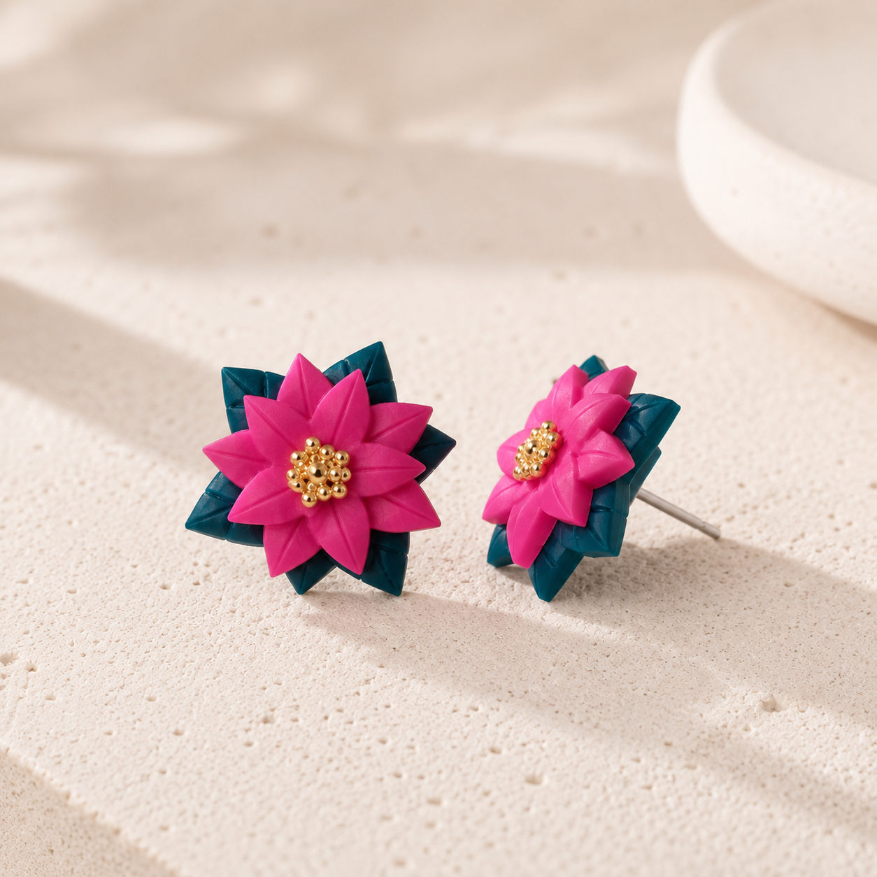A pair of flower-shaped stud earrings with bright pink petals, dark teal layered leaves, and a small gold bead center, displayed on a textured beige surface.