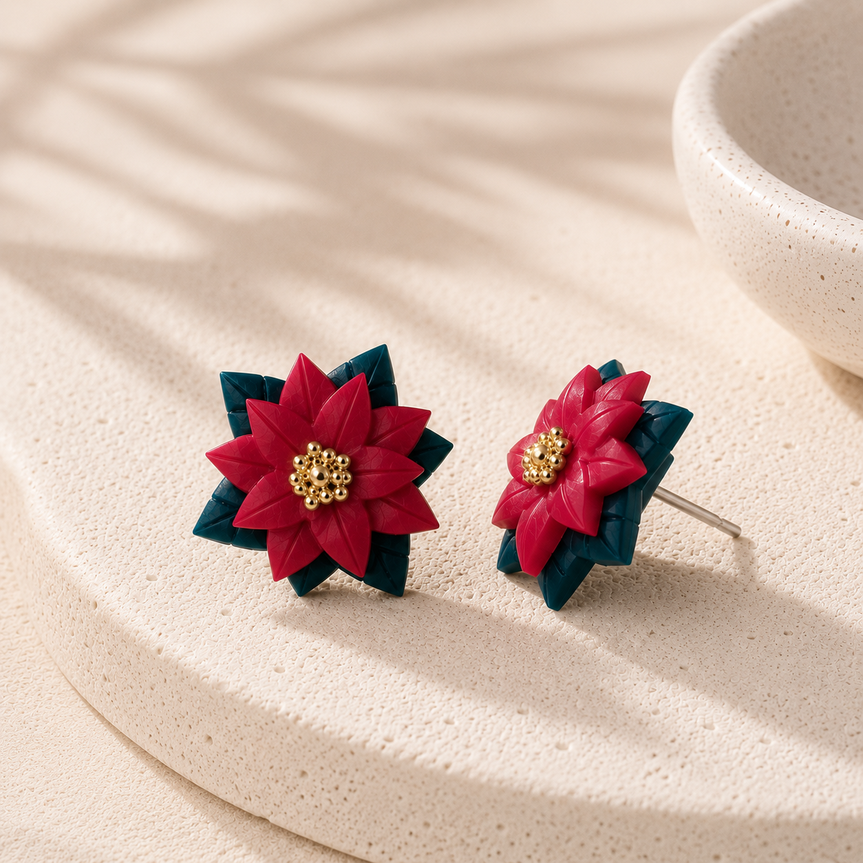 A pair of handmade flower stud earrings with dark red petals, dark teal layered leaves, and a gold beaded center, displayed on a textured beige stone surface with soft leaf shadows.
