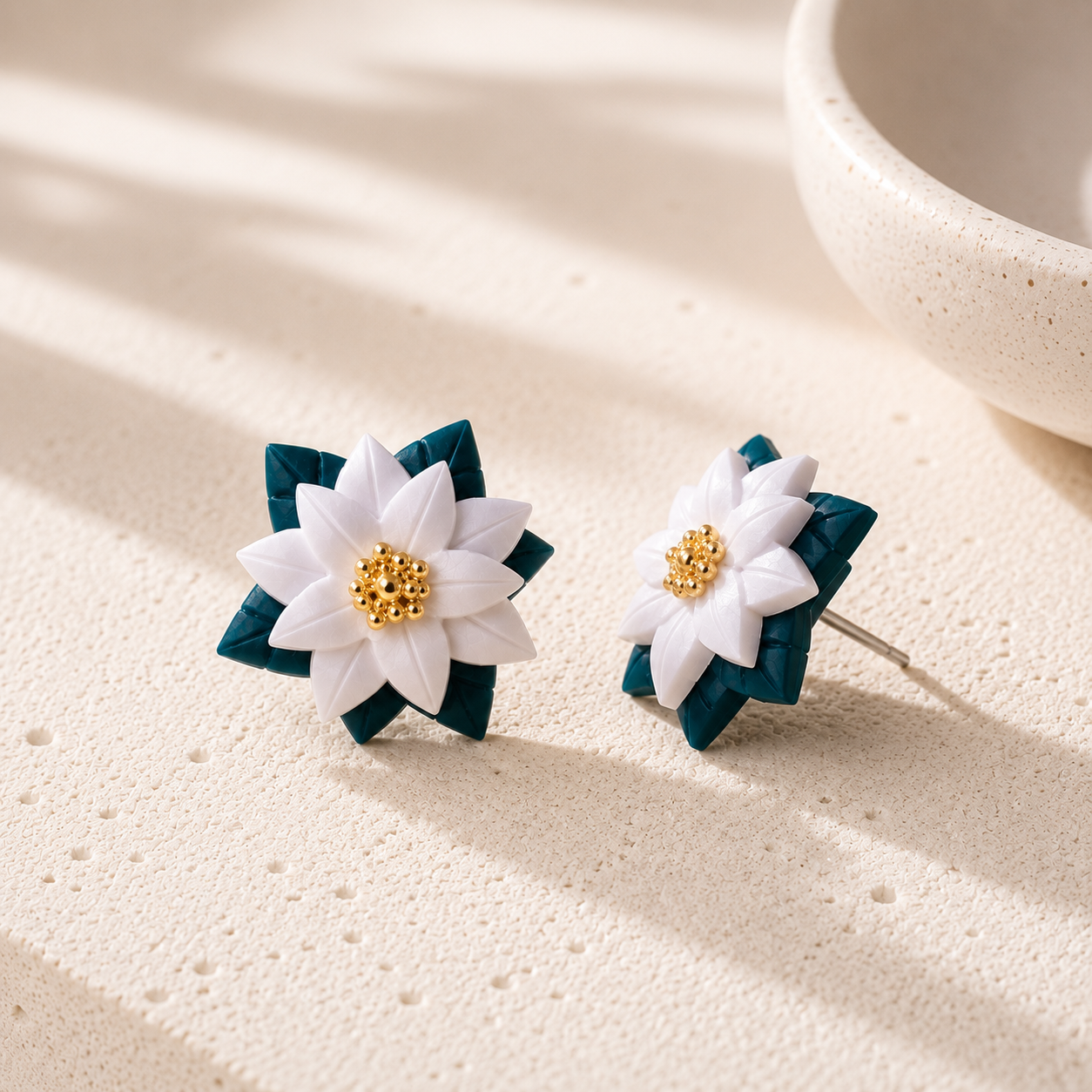A pair of handmade flower stud earrings with white petals, dark teal layered leaves, and a gold beaded center, displayed on a textured beige surface with soft shadows.