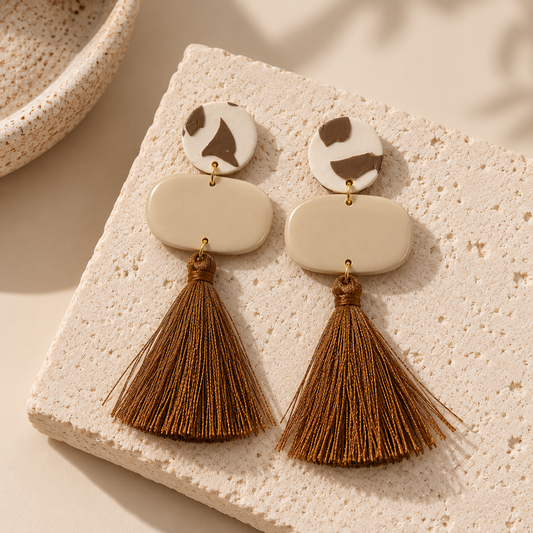 A pair of bohemian-style drop earrings with cream-colored geometric discs, brown abstract accents on the top discs, and long brown tassel charms, displayed on a textured beige stone block.
