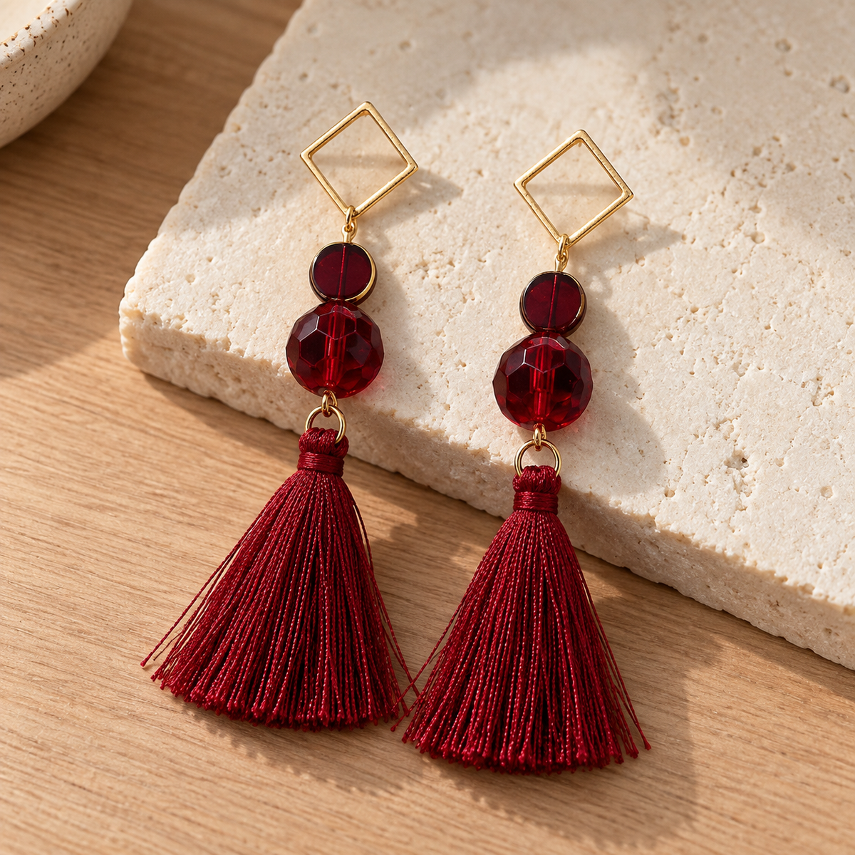 A pair of gold-tone statement earrings with deep red faceted beads, square post tops, and long red tassel drops, displayed on a textured beige stone slab over a light wood surface.