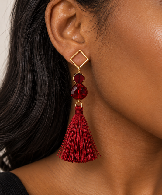 A close-up of a model's ear wearing a dangling earring with a gold diamond-shaped stud, a round red gemstone, a faceted red bead, and a long red tassel.