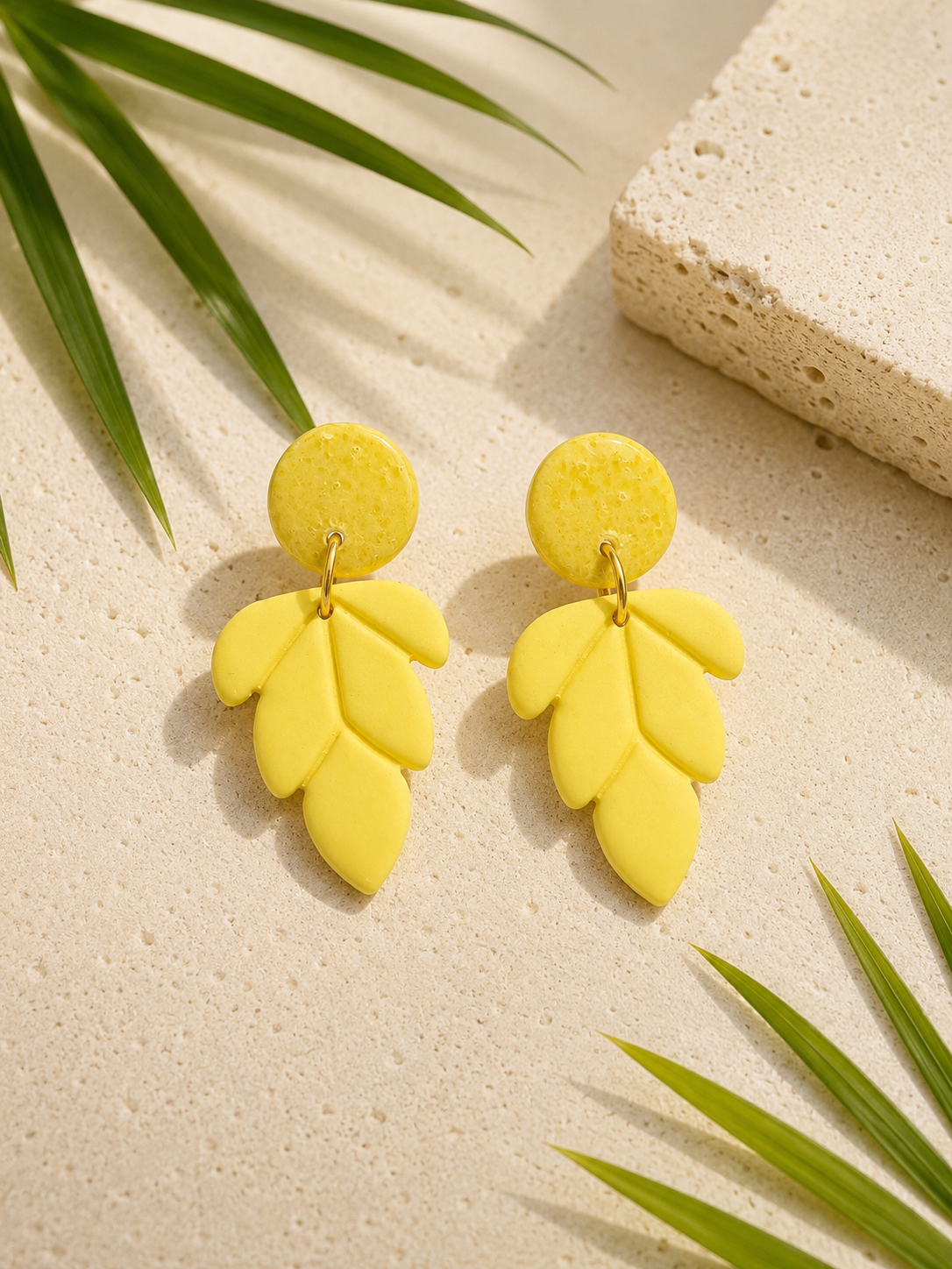 Yellow leaf-shaped earrings on a beige surface with green leaves.