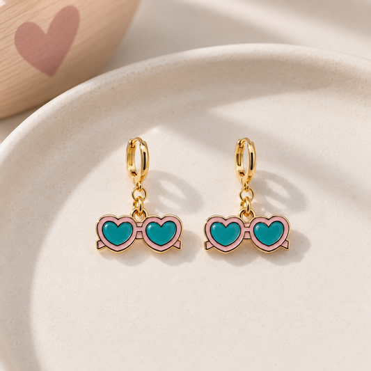 A pair of gold hoop earrings with dangling heart-shaped glasses charms featuring turquoise blue lenses and pink frames, displayed on a light beige ceramic dish.