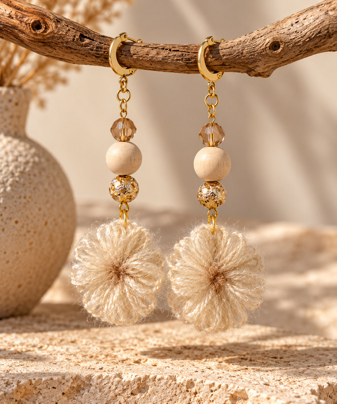 Boho-style dangle earrings with gold-tone hoops, wooden beads, clear faceted beads, and fluffy cream pom-poms, hanging from a rustic branch in a warm neutral setting.