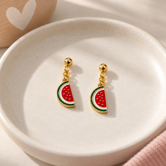 A pair of gold-tone dangle earrings shaped like watermelon slices, resting on a small round beige dish. The earrings feature red watermelon flesh with green and white rind details.
